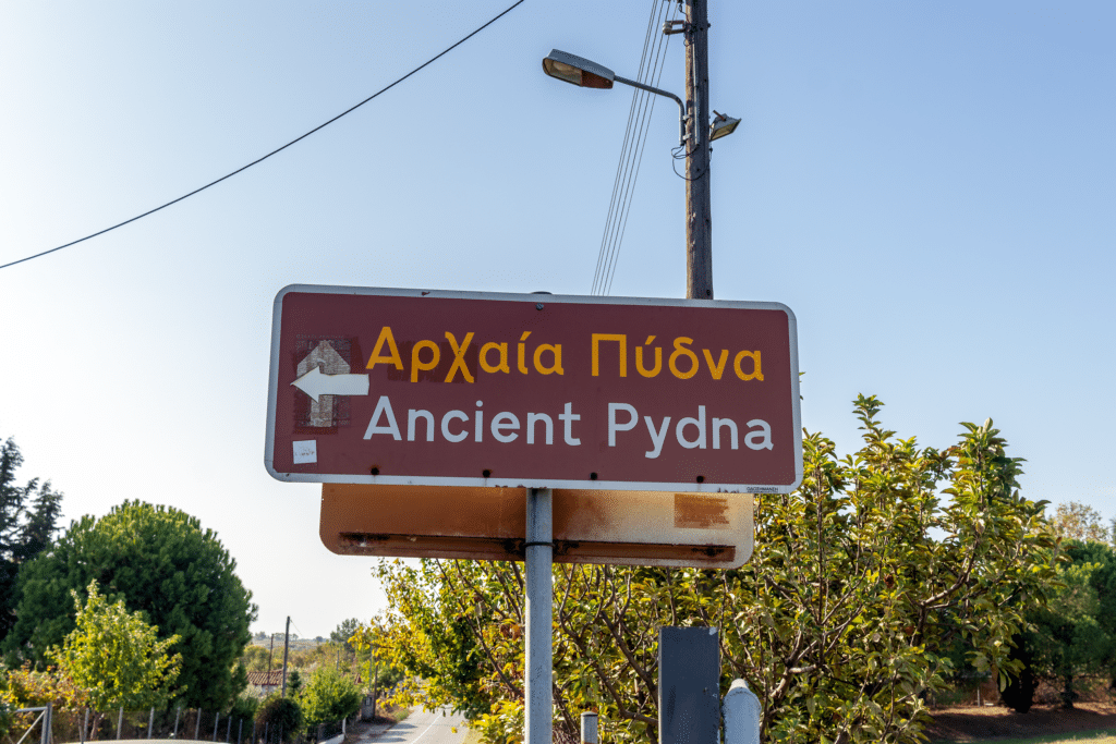 red road sign with the word of Ancient Pydna in English and Greek in Pieria region with an arrow pointed to the left.